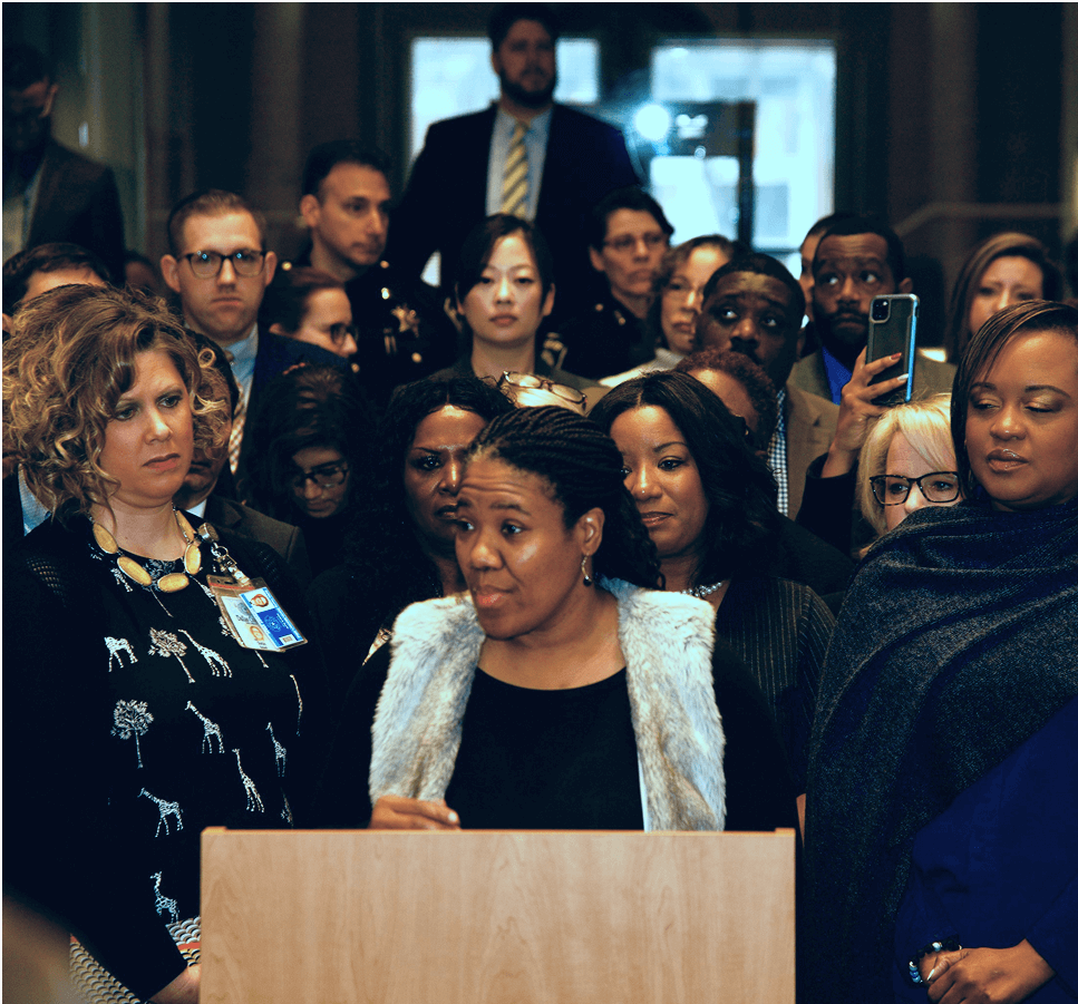 img8 Tonya speaking at a podium with a large group of people behind her.